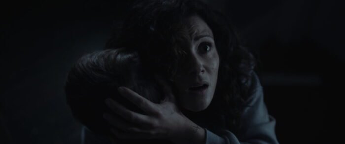 A woman holds her son in her arms while looking up toward the camera in horror (The Confession)