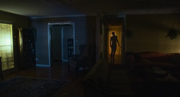 A dark room is accentuated by a silhouette emerging from the light of a room at the end of a hallway. (The Other People)