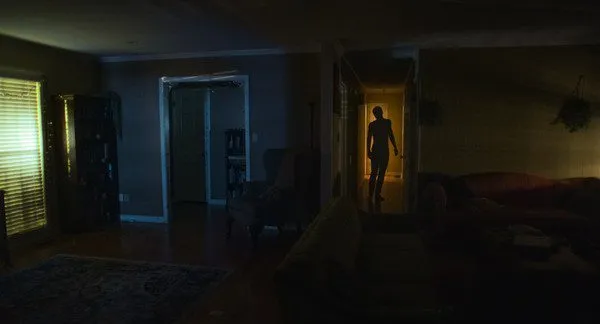 A dark room is accentuated by a silhouette emerging from the light of a room at the end of a hallway. (The Other People)