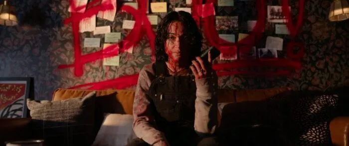 A woman with a bloody face sits on the edge of a bed holding a phone in her hand. A spray painted wall is behind her. (The Red Mask / FrightFest)