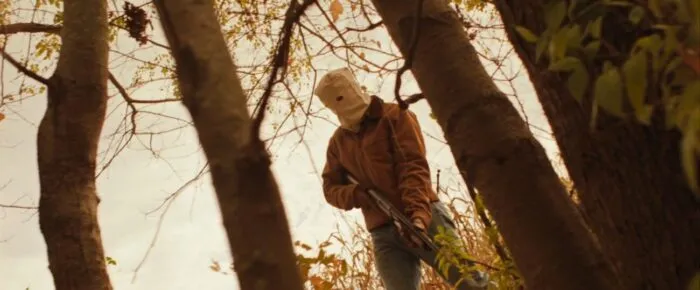A masked person holding a shotgun in the woods (The Rows)