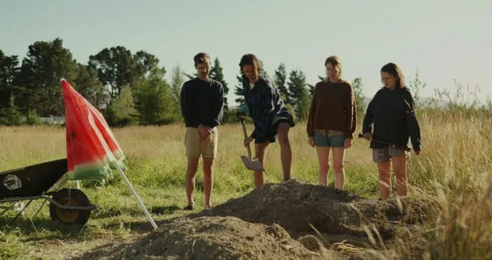 three onlookers watch a woman dig a hole in a field (The Weed Eaters)