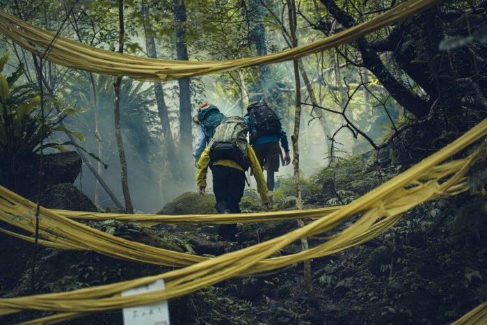 Three people hike past hanging yellow fabric that resembles caution tape. (Haunted Mountains: The Yellow Taboo)