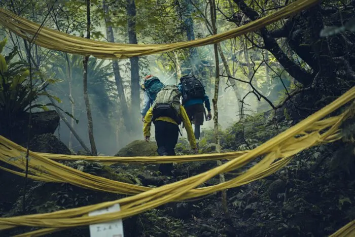 Three people hike past hanging yellow fabric that resembles caution tape. (Haunted Mountains: The Yellow Taboo)