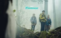 something in the foreground catches the attention of three individuals standing on a high rock in the fog of a forest.