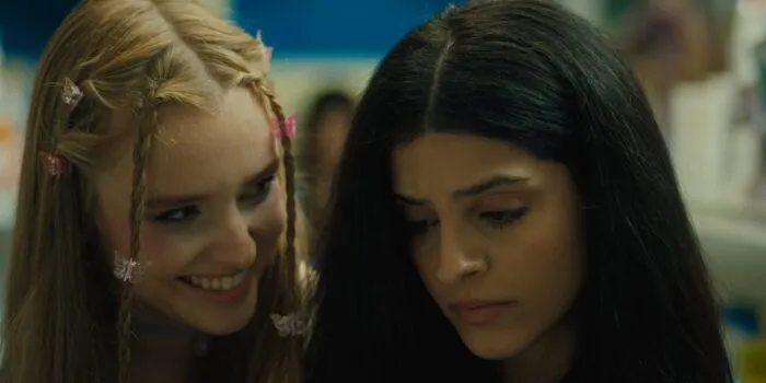 A young blonde woman in braids smiles devilishly inside the personal space of a less outgoing Yasamin. (Foreigner)