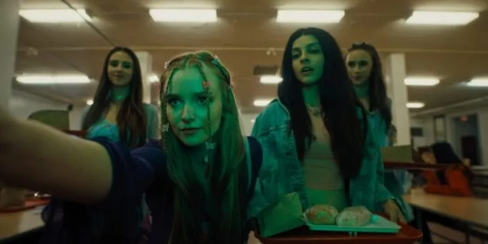 A young woman with braids and butterfly clips outstretches her arm past the camera while three other girls stand behind her, one holding a lunch tray.