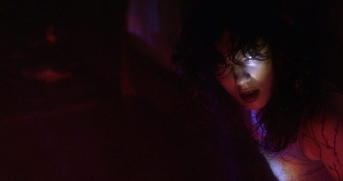 A woman in low light hides tightly against a bed.