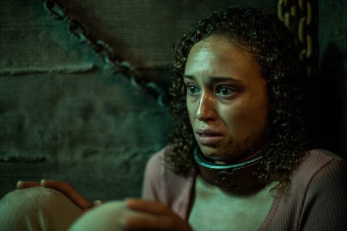 A woman is seen chained by the neck while sitting with her back to the wall