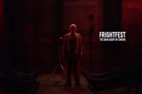 A man stands in front of a camera under a dark red light between the feet of two bodies lying on the ground