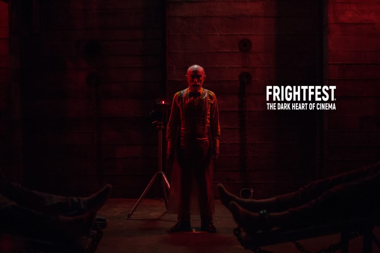 A man stands in front of a camera under a dark red light between the feet of two bodies lying on the ground