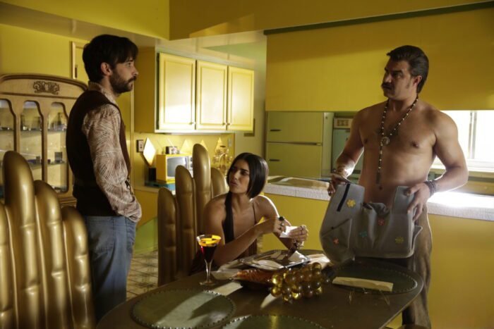 two men standing and a woman sitting down around a kitchen table.