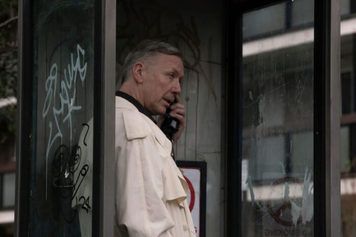 A man in a white trench coat talks on a payphone.