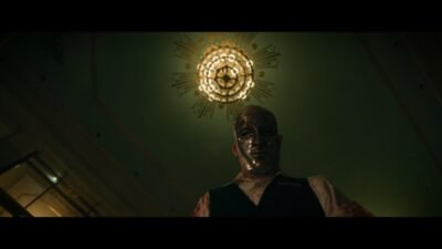 A well-dressed masked man stands under a chandelier and gazes into the camera.