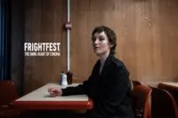 A woman with her makeup running down her cheeks sits at an old restaurant table against a wall with wood paneling (Odyssey, 2025)