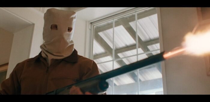 A hooded man blasts a shotgun inside a house