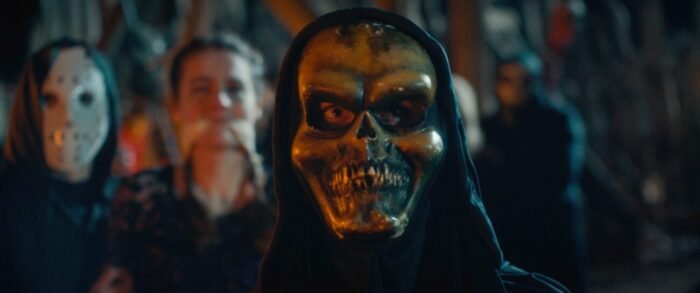 A masked and cloaked person stands in front of a woman being gagged and held by another person in a mask and cloak.