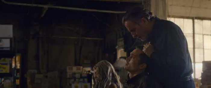 Nico stands over John, who is seated in a chair, pulling his hair and looking down into his eyes while speaking to him. Edie sits in a shair int the background.