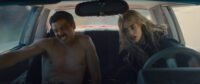 John is seen naked in the passenger seat while Edie is seen driving a car, both are leaning to their right as the car turns. (Eeenie Meanie)