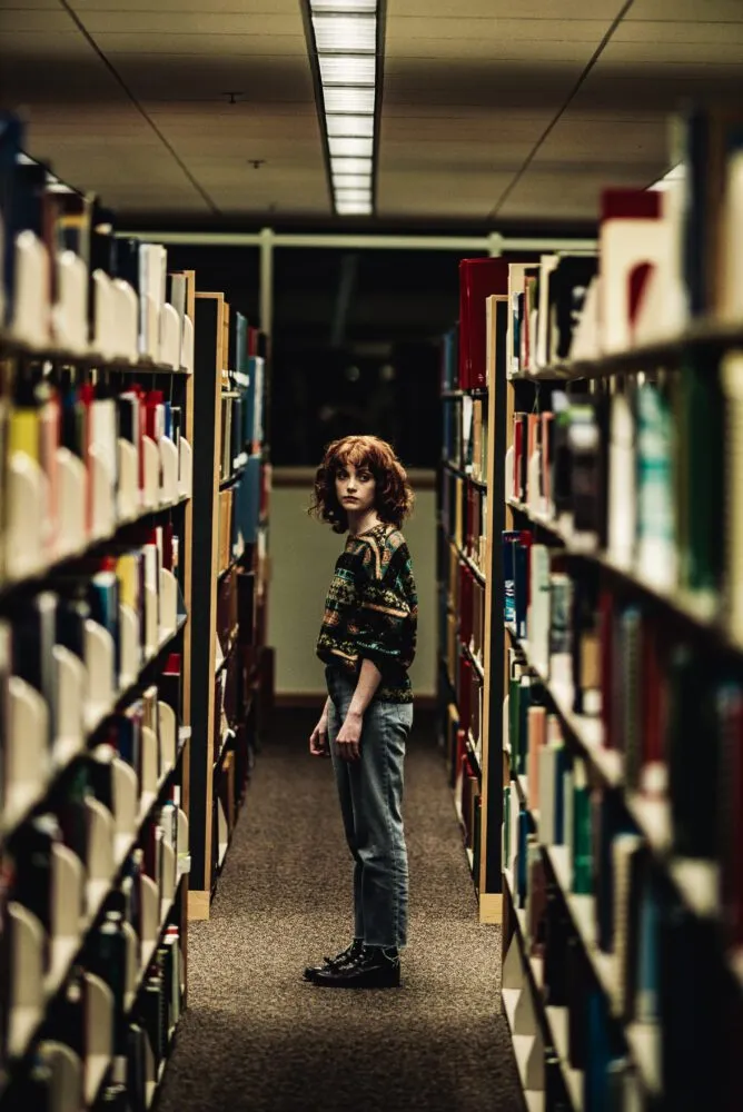 A woman with red hair in a sweater stands between rows of books in a library.