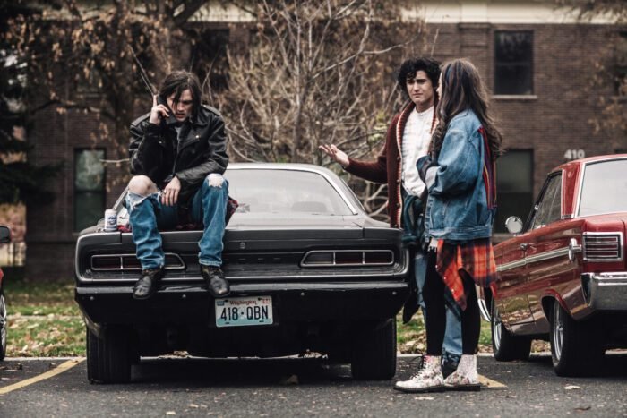 A man speaks on an early cell phone while sitting on the trunk of his car while a man and woman look at each other confused.