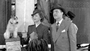 Man in a double breasted coat and hat standing next to a woman holding a coat. Next to them is a small dog looking at them, they are looking past the camera.