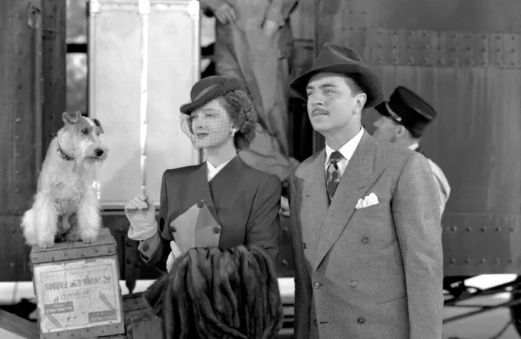 Man in a double breasted coat and hat standing next to a woman holding a coat. Next to them is a small dog looking at them, they are looking past the camera.