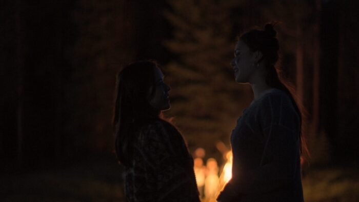 two people shrouded in darkness are silhouetted by the bonfire in the background.