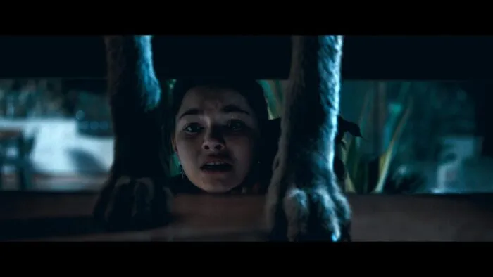 A young girl holds her dog underneath a staircase as she watches a coyote walk down the stairs. 