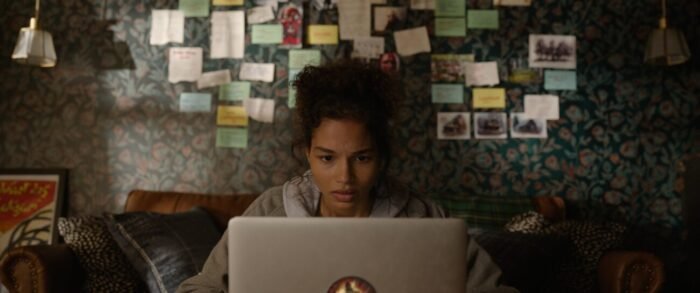 Allina is seen typing on her computer in front of a wall pinned with notes.