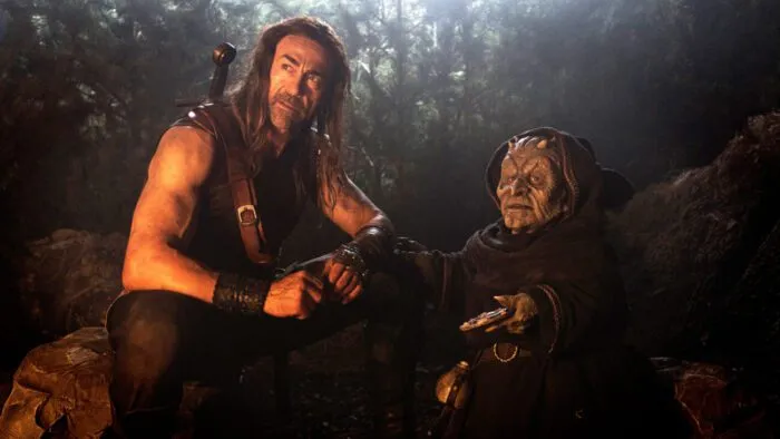 a muscular man with long hair and a sword on his back speaks with a goblin by a fire.