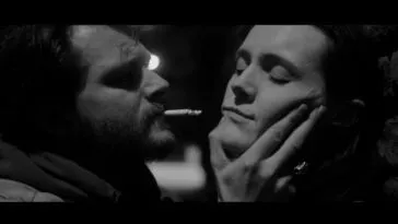 A man with a cigarette in his mouth gets uncomfortably close to a woman he's holding by the jaw.