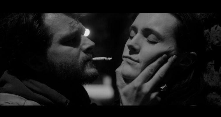 A man with a cigarette in his mouth gets uncomfortably close to a woman he's holding by the jaw.