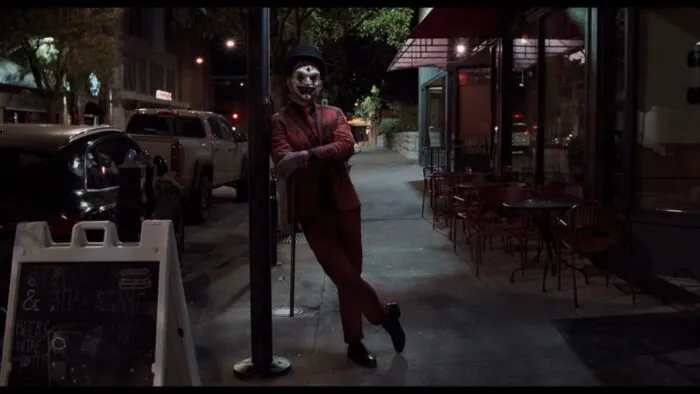 The Jester leans up against a street light on a busy sidewalk.