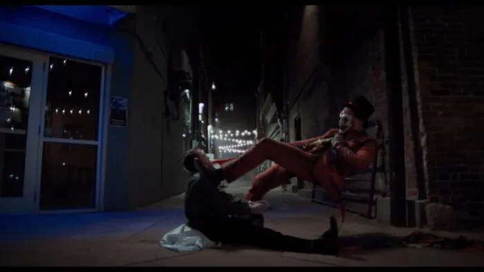 The Jester uses his cane as leverage while pulling on a handkerchief rope from a man laying down in an alley.