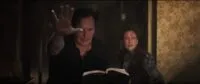 a man with his hand extended reading from a book and a scared and worried woman over his shoulder.