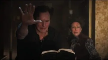 a man with his hand extended reading from a book and a scared and worried woman over his shoulder.