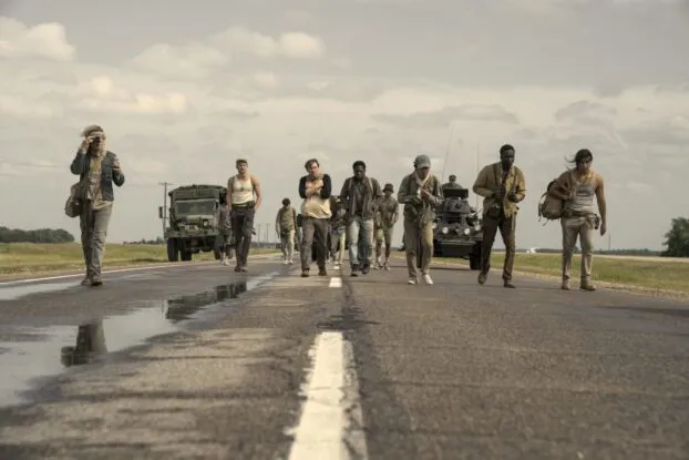 The boys walk down a road in the summer heat. A few vehicles follow closely behind them.