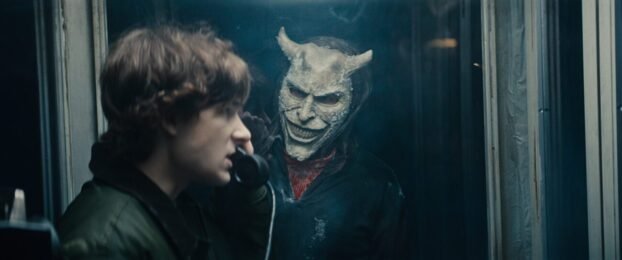 Finney stands in a phone booth with the Black Phone in his hand, while the Grabber stands outside looking at him.