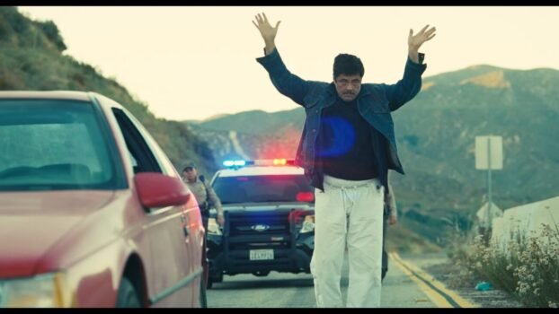 Sergio St. Carlos (Benicio del Toro) stands by his car with a police car in the background. His hands are in the air.