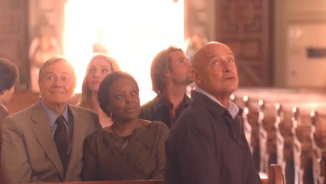 A ground of people sit in church pews and look around, behind them a bright light shines through the doors