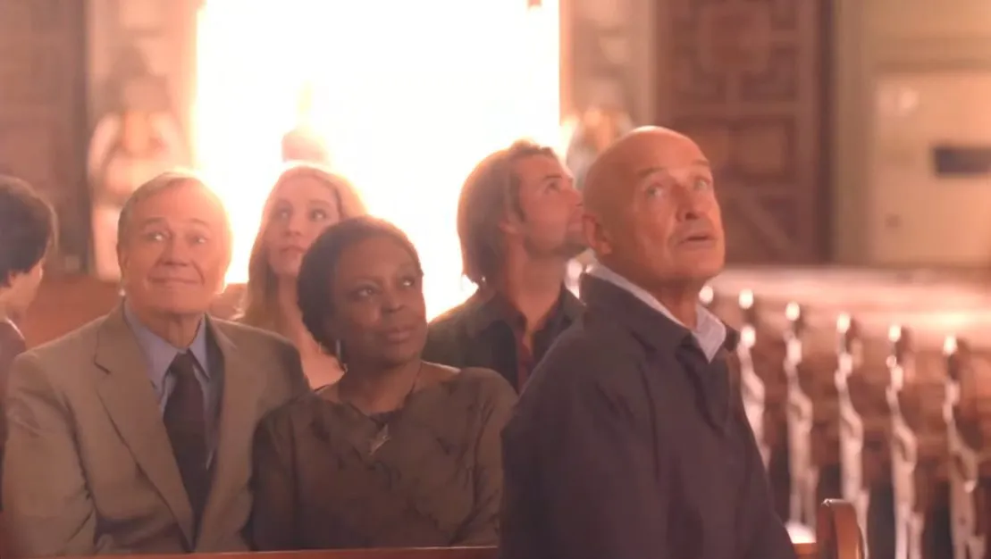 A ground of people sit in church pews and look around, behind them a bright light shines through the doors