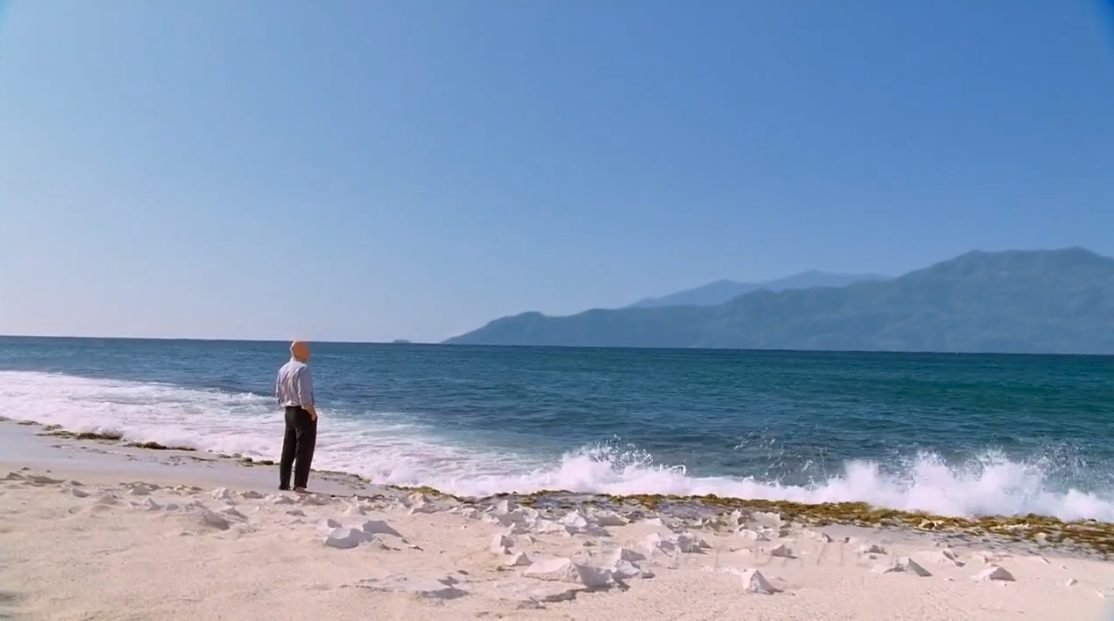 An older man in black pants and a blue shirt stands in the sand and looks out over the ocean to an island in the distance