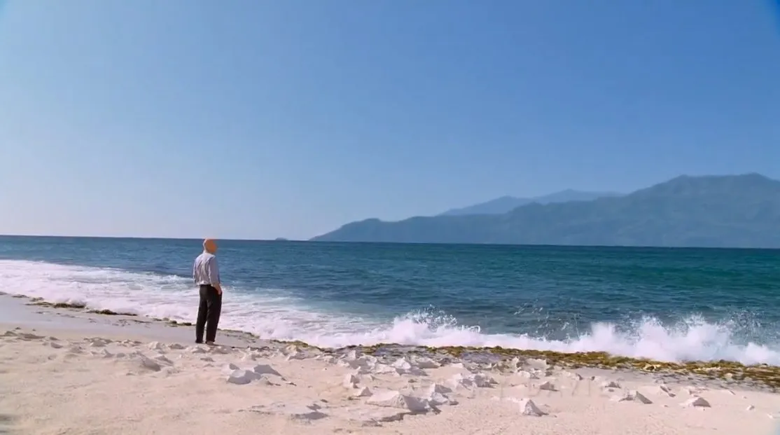 An older man in black pants and a blue shirt stands in the sand and looks out over the ocean to an island in the distance