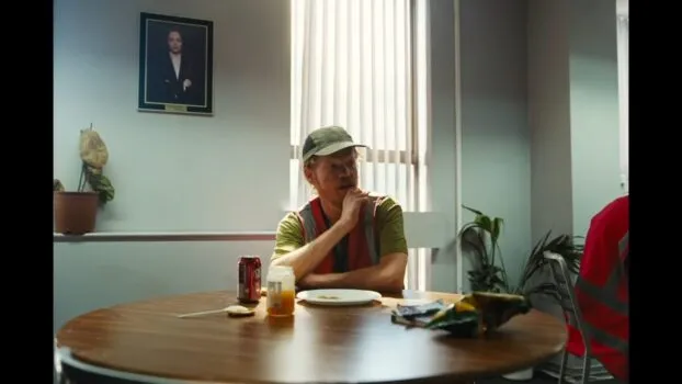 Teddy (Jesse Plemons) sits at a table in his work uniform eating lunch and watching TV.