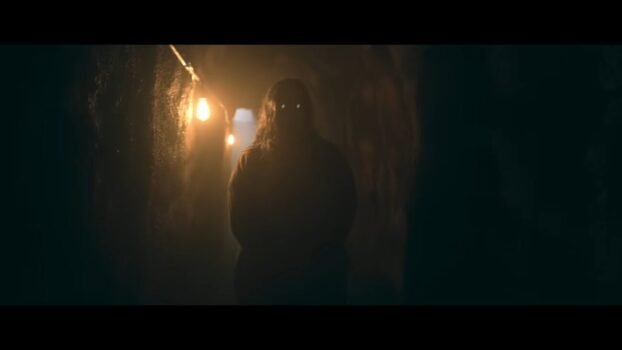 An old lady stands in an underground hallway with glowing eyes.