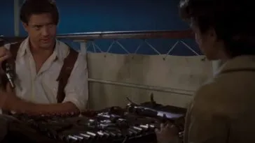 A man in a white shirt places a handgun in a shoulder holster, and looks across a table at a woman with her back to us. They are both sitting by the rail of a boat