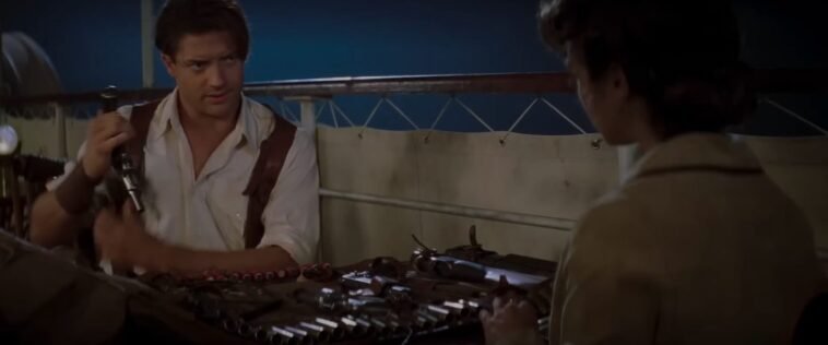 A man in a white shirt places a handgun in a shoulder holster, and looks across a table at a woman with her back to us. They are both sitting by the rail of a boat