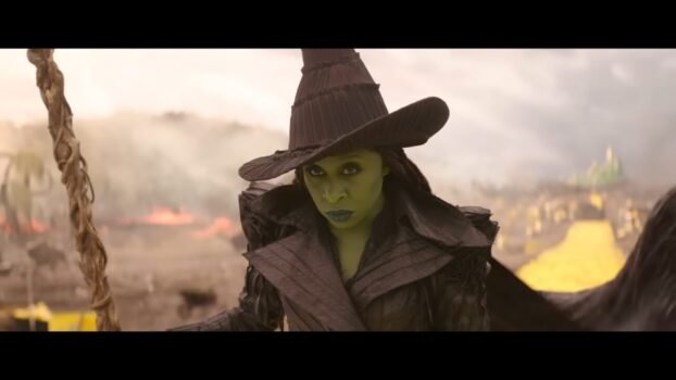 Elphaba (Cynthia Erivo) lands on the Yellow Brick Road with her broom in hand. She is wearing black with a hat and matching outfit.