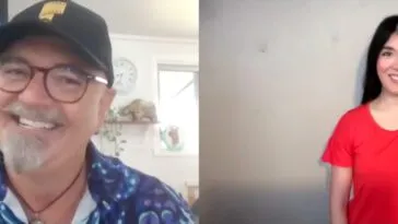 Neil Fanning, wearing a black cap and blue camp shirt with a poster for “Scooby-Doo” (2002) in the background, smiles while talking to Jamie Lee, wearing an orange shirt, in an interview.